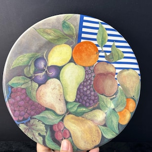 May include: Round decorative plate featuring a still life of various fruits, including pears, grapes, oranges, and lemons. The plate has a gray background with a blue and white striped section. Green leaves are scattered throughout the design.