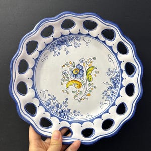 May include: A white ceramic plate with a blue floral design and a pierced edge. The plate has a scalloped rim and oval cutouts. The central design is a hand-painted floral motif. The rim is painted blue. The plate is held by a hand.