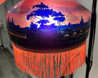 May include: A decorative lamp with a painted glass shade featuring a landscape scene with a tree and buildings. The shade has a gradient of colors, including red, blue, and purple. The lamp has a fringe trim in orange.