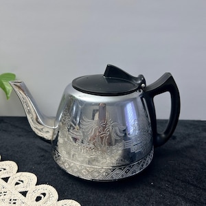 May include: A silver teapot with a black handle and lid. The teapot features an etched design around the body. The spout is curved, and the handle is a simple, curved design. A classic kitchenware item, the teapot is a stylish addition.
