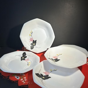 Peut inclure: Ensemble de quatre assiettes octogonales blanches avec un motif floral. Chaque assiette présente une fleur noire avec des accents roses et jaunes. Les assiettes sont disposées sur une nappe rouge.
