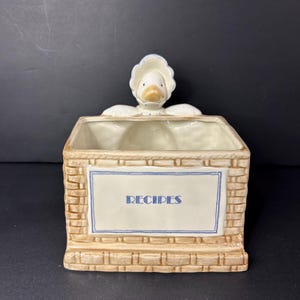 May include: A ceramic recipe box featuring a duck figurine. The box has a rectangular shape with a brickwork design and a label with the word "RECIPES". The duck is wearing a bonnet.