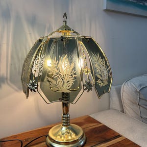 Pode incluir: Uma luminária de mesa dourada com um design floral na cúpula. A luminária tem uma base redonda e uma haste, e a cúpula tem a forma de uma flor. A luminária está em uma mesa de madeira.