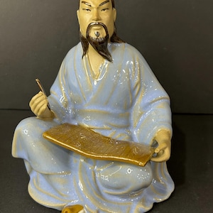 Op de afbeelding: Een keramieken beeldje van een zittende Aziatische man in een lichtblauwe jurk, met een schrijfinstrument en een rol. De figuur heeft een lange zwarte baard en draagt een gele hoofdtooi. Een decoratief item.