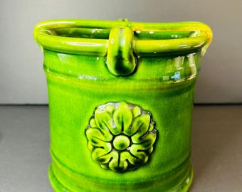 May include: A green ceramic planter with a flower design. The planter has a handle on each side and a rounded bottom.