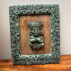 May include: A framed relief sculpture featuring a teal and cream-colored frame with intricate carvings. The sculpture depicts a figure with a headdress and ornate details, set against a wooden background. The artwork is displayed on a wooden surface.