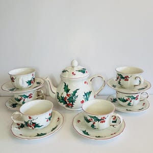 Puede incluir: Un juego de té de cerámica blanca con una tetera, seis tazas de té y platillos. El juego está decorado con un diseño de acebo y bayas en verde y rojo, con un borde rojo. La tetera tiene tapa y boquilla curva.