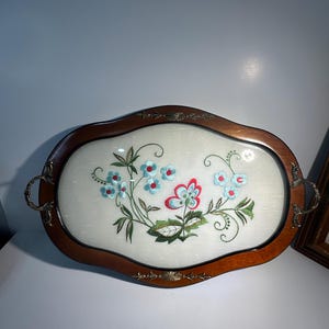 May include: Oval serving tray with a wooden frame and a cream-coloured centre. The tray features embroidered flowers in blue, red, and green. It has metal handles and trim.