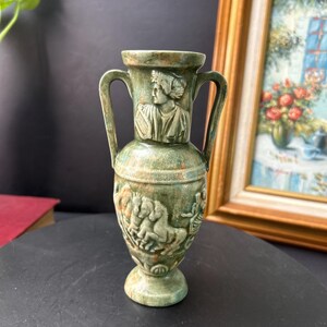 May include: A small, green ceramic vase with two handles. The vase features relief carvings of a classical figure and horses. The vase has a weathered appearance, suggesting an antique or vintage aesthetic. The vase is displayed on a dark surface.