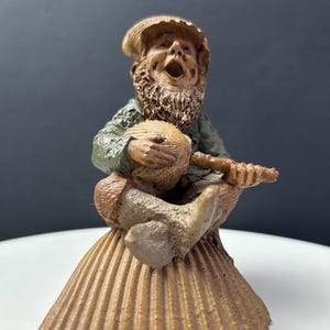 May include: A detailed figurine of a bearded man playing a stringed instrument. The figure is dressed in a green jacket and hat, seated on a brown, shell-shaped base. The man's mouth is open as if he is singing or speaking.