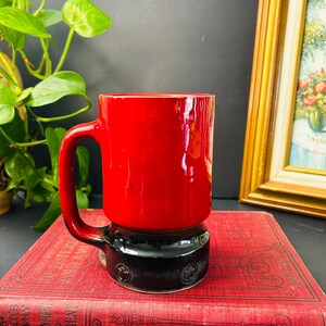Peut inclure: Une tasse en céramique rouge et noire avec une anse. La partie supérieure de la tasse est rouge brillant, tandis que la base est noire avec des motifs circulaires en relief. La tasse est posée sur un livre rouge.