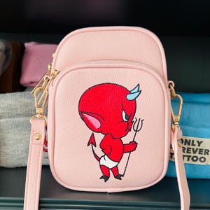 Puede incluir: Un bolso bandolera de color rosa claro con un gráfico de un personaje de diablo rojo con cuernos azules que sostiene un tridente. El bolso tiene un compartimento principal con cremallera, un bolsillo frontal con cremallera y herrajes dorados.