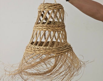Pode incluir: Uma luminária pendente tecida em tons naturais, com base alargada e alça trançada. A luminária tem um design em camadas e uma borda com franjas. A luminária é segurada por uma mão em um fundo branco.