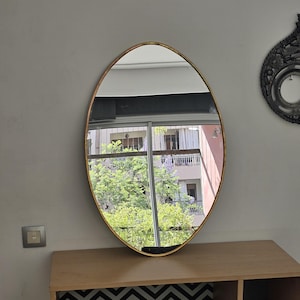 May include: A gold oval-shaped mirror with a beveled edge hangs on a white wall above a wooden shelf with black and white chevron patterned paper lining the back of the shelf.