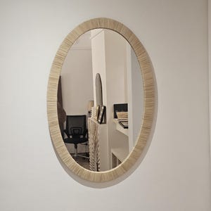 May include: Oval mirror with a woven, light-colored frame. The mirror is hanging on a white wall, reflecting a room with a black chair and other furnishings. The frame has a textured appearance.