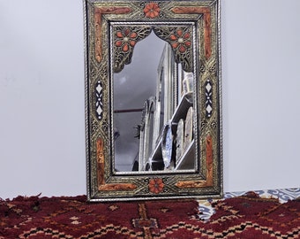 May include: A silver-framed mirror with intricate carvings and red and gold accents. The mirror has an arched top and is reflecting other mirrors and decorative items.