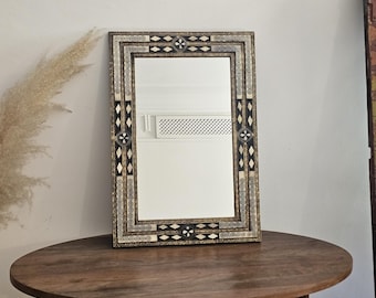 May include: A rectangular wall mirror featuring an intricate frame with alternating black and cream-colored geometric inlays, including diamond shapes and circular patterns, surrounding a clear, reflective glass center panel.