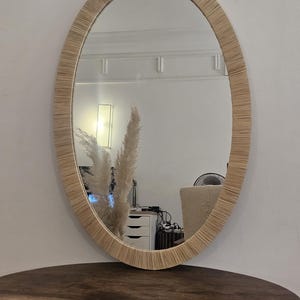 May include: Oval mirror with a woven, light brown frame. The mirror is hanging on a white wall and reflects a room with a table, a lamp, and decorative plants. The mirror is a decorative home decor item.