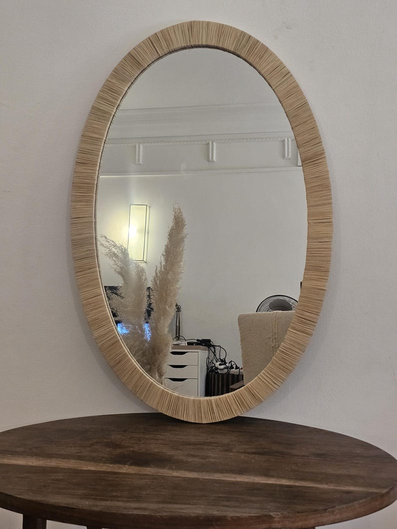 May include: Oval mirror with a woven, light brown frame. The mirror is hanging on a white wall and reflects a room with a table, a lamp, and decorative plants. The mirror is a decorative home decor item.