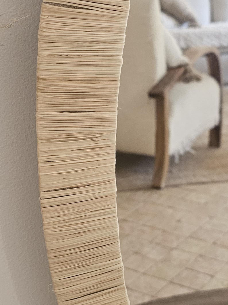 May include: A round mirror with a natural woven frame. The frame is made of tightly woven, light-colored fibers, creating a textured surface. The mirror is mounted on a white wall, with a blurred background showing a glimpse of a room.