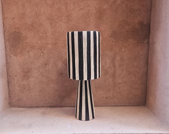 May include: A black and white striped table lamp with a conical base. The lamp is sitting on a shelf against a textured wall.
