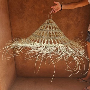May include: A handcrafted, natural straw sun hat with a conical shape and a wide, fringed brim. The hat is made of woven straw and has a loop at the top for hanging. The hat is held up against a reddish-brown wall.