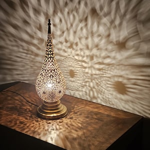 May include: A decorative brass table lamp with a pierced, teardrop-shaped shade. The lamp casts intricate patterns of light onto the wall and wooden surface below. The lamp has a round base and a tall, pointed top.