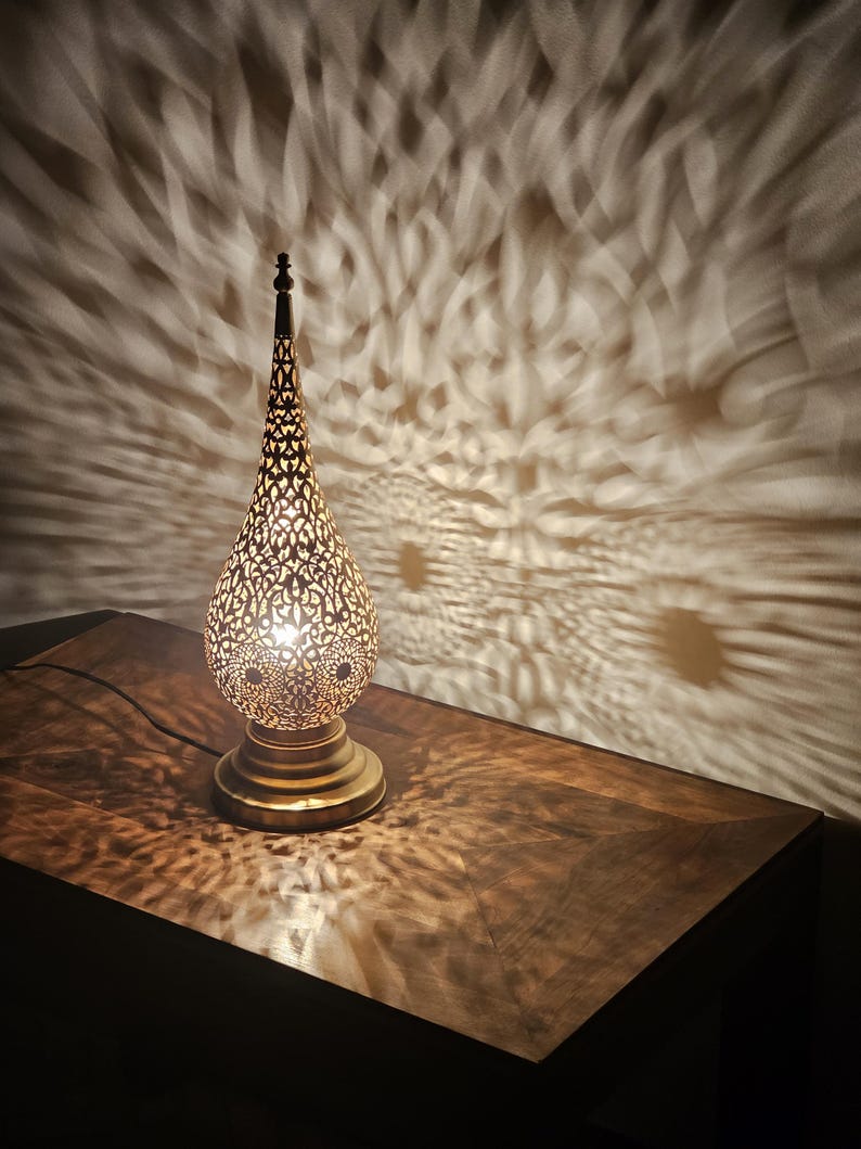 May include: A decorative brass table lamp with a pierced, teardrop-shaped shade. The lamp casts intricate patterns of light onto the wall and wooden surface below. The lamp has a round base and a tall, pointed top.