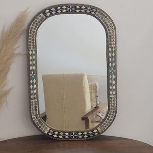 Puede incluir: Espejo ovalado con marco decorativo. El marco presenta un patrón de formas geométricas en blanco y negro y diseños circulares. El espejo refleja una silla y una pared de color neutro. El espejo está sobre una mesa de madera oscura.