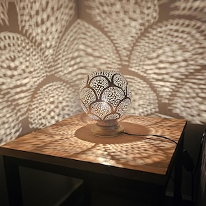 Puede incluir: Una l&aacute;mpara de mesa redonda de metal con un dise&ntilde;o decorativo recortado. La l&aacute;mpara est&aacute; sobre una mesa de madera y proyecta sombras intrincadas en la superficie.