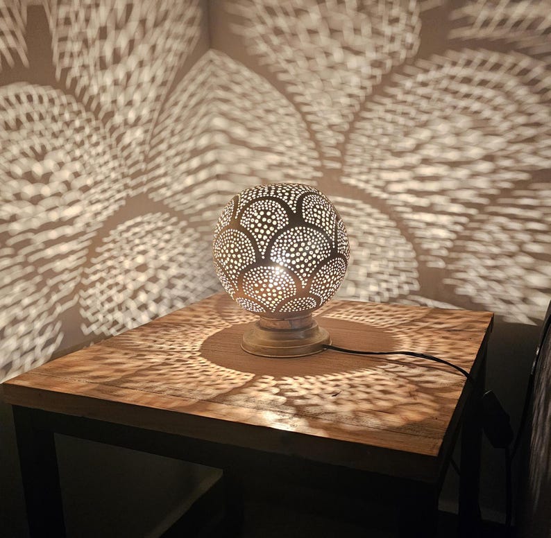 Puede incluir: Una l&aacute;mpara de mesa redonda de metal con un dise&ntilde;o decorativo recortado. La l&aacute;mpara est&aacute; sobre una mesa de madera y proyecta sombras intrincadas en la superficie.