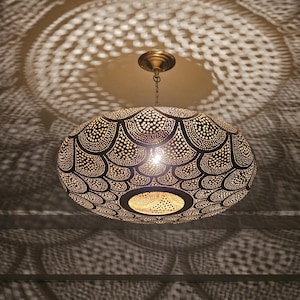 Pode incluir: Uma luminária pendente ovalada e ornamentada, com um design perfurado detalhado. A luz projeta sombras intrincadas. Suspensa por uma corrente, emite um brilho quente. O design cria um efeito decorativo no teto.