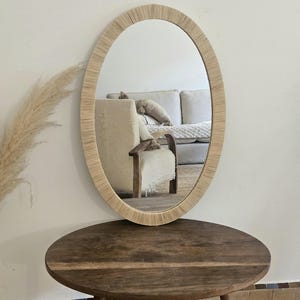 May include: Oval mirror with a woven, light-colored frame. The mirror is displayed on a dark brown, round wooden table with three legs. The reflection shows a neutral-toned living room.