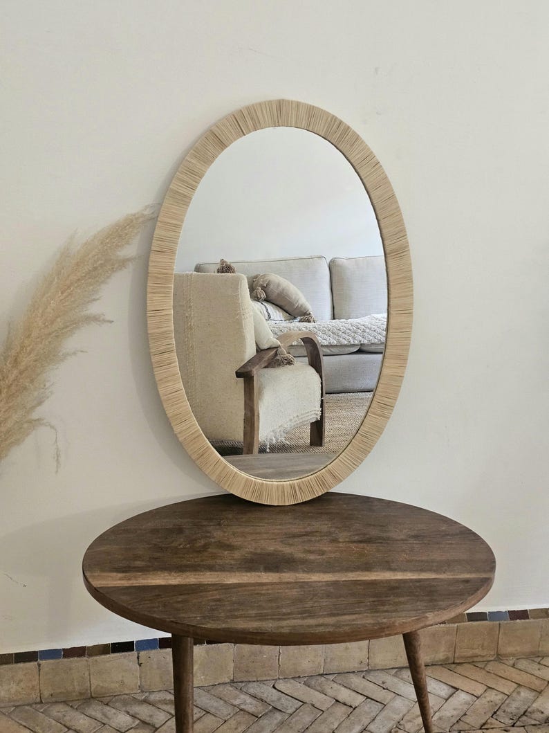 May include: Oval mirror with a woven, light-colored frame. The mirror is displayed on a dark brown, round wooden table with three legs. The reflection shows a neutral-toned living room.