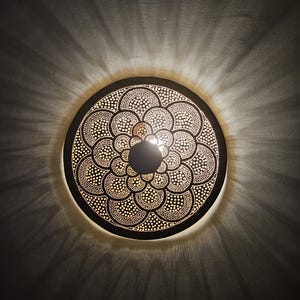 Op de afbeelding: Een ronde, decoratieve wandlamp met een gedetailleerd, geperforeerd ontwerp. De lamp is gemaakt van donker metaal en heeft een patroon van kleine, gelijkmatig verdeelde stippen. Het licht werpt een radiaal patroon op de omringende muur.