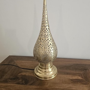 May include: A gold-colored metal table lamp with a decorative, pierced design. The lamp has a teardrop-shaped shade with intricate cut-out patterns and a round base. The lamp is on a wooden surface.
