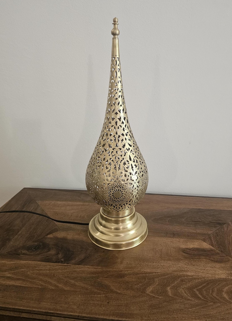 May include: A gold-colored metal table lamp with a decorative, pierced design. The lamp has a teardrop-shaped shade with intricate cut-out patterns and a round base. The lamp is on a wooden surface.