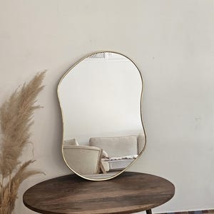 May include: An irregularly shaped mirror with a gold-colored frame. The mirror is displayed on a dark brown, oval-shaped wooden table with three legs. The reflection shows a glimpse of a room with a sofa and a white wall.