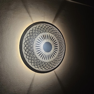 May include: A round, black wall sconce with a geometric design. The sconce is lit and casting a warm glow on the wall.