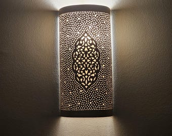 May include: A cylindrical wall sconce with a perforated design, emitting a warm glow. The lamp features a central, ornate cutout pattern. The light fixture is mounted on a textured wall, creating a play of light and shadow.