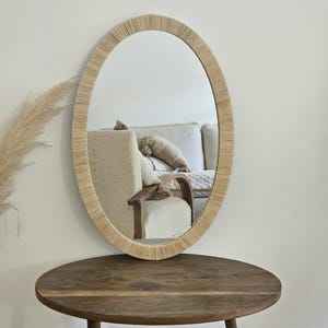 May include: Oval mirror with a woven, light-colored frame. The mirror hangs on a white wall above a round, wooden table. The reflection shows a glimpse of a beige sofa and chair.