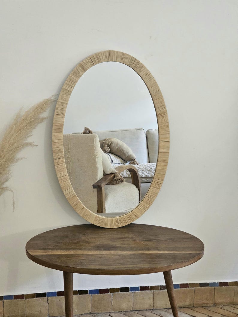 May include: Oval mirror with a woven, light-colored frame. The mirror hangs on a white wall above a round, wooden table. The reflection shows a glimpse of a beige sofa and chair.