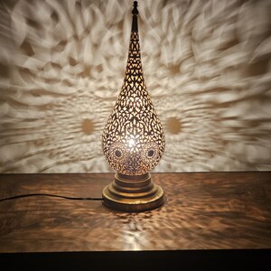 May include: A decorative brass table lamp with an intricate, openwork design. The lamp has a teardrop-shaped shade and a round base. The light from the lamp casts a patterned shadow on the wall behind it, creating a warm and inviting ambiance.