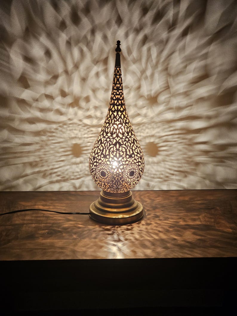 May include: A decorative brass table lamp with an intricate, openwork design. The lamp has a teardrop-shaped shade and a round base. The light from the lamp casts a patterned shadow on the wall behind it, creating a warm and inviting ambiance.