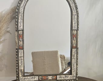 May include: An arched mirror with an ornate frame. The frame features a cream-colored base with intricate black and orange geometric patterns. The mirror is displayed on a round, dark wooden table, reflecting a neutral-toned chair.