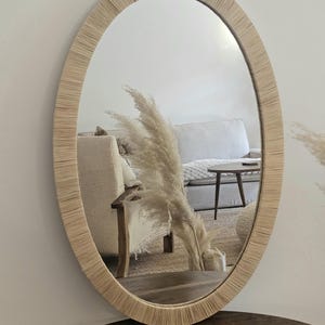 May include: Oval mirror with a natural woven frame. The mirror reflects a living room scene with a sofa, table, and decorative pampas grass. The frame has a light beige color, adding a touch of rustic charm to the decor.