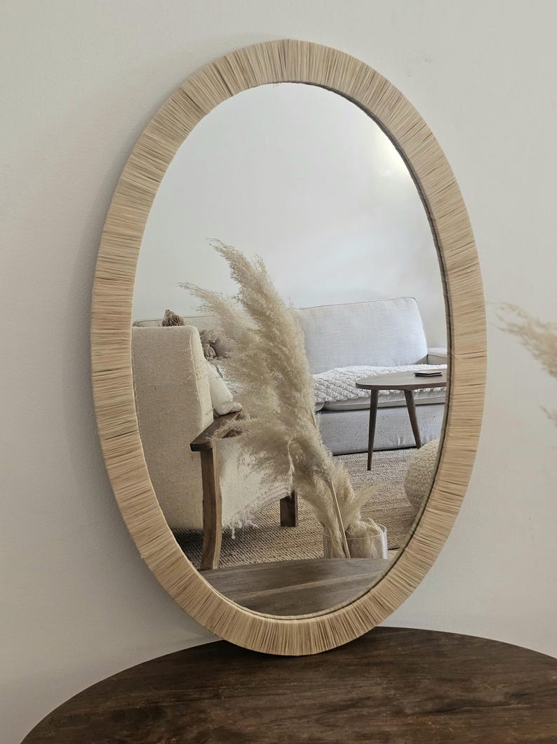 May include: Oval mirror with a natural woven frame. The mirror reflects a living room scene with a sofa, table, and decorative pampas grass. The frame has a light beige color, adding a touch of rustic charm to the decor.