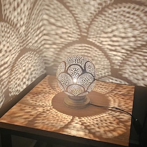 Puede incluir: Una l&aacute;mpara de mesa plateada con una pantalla redonda y estampada proyecta intrincadas sombras geom&eacute;tricas sobre una mesa de madera.