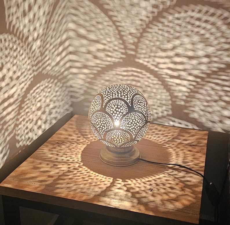 Puede incluir: Una l&aacute;mpara de mesa plateada con una pantalla redonda y estampada proyecta intrincadas sombras geom&eacute;tricas sobre una mesa de madera.
