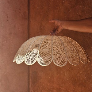 May include: A white crocheted lampshade with a scalloped edge hangs from a hand against a brown wall.